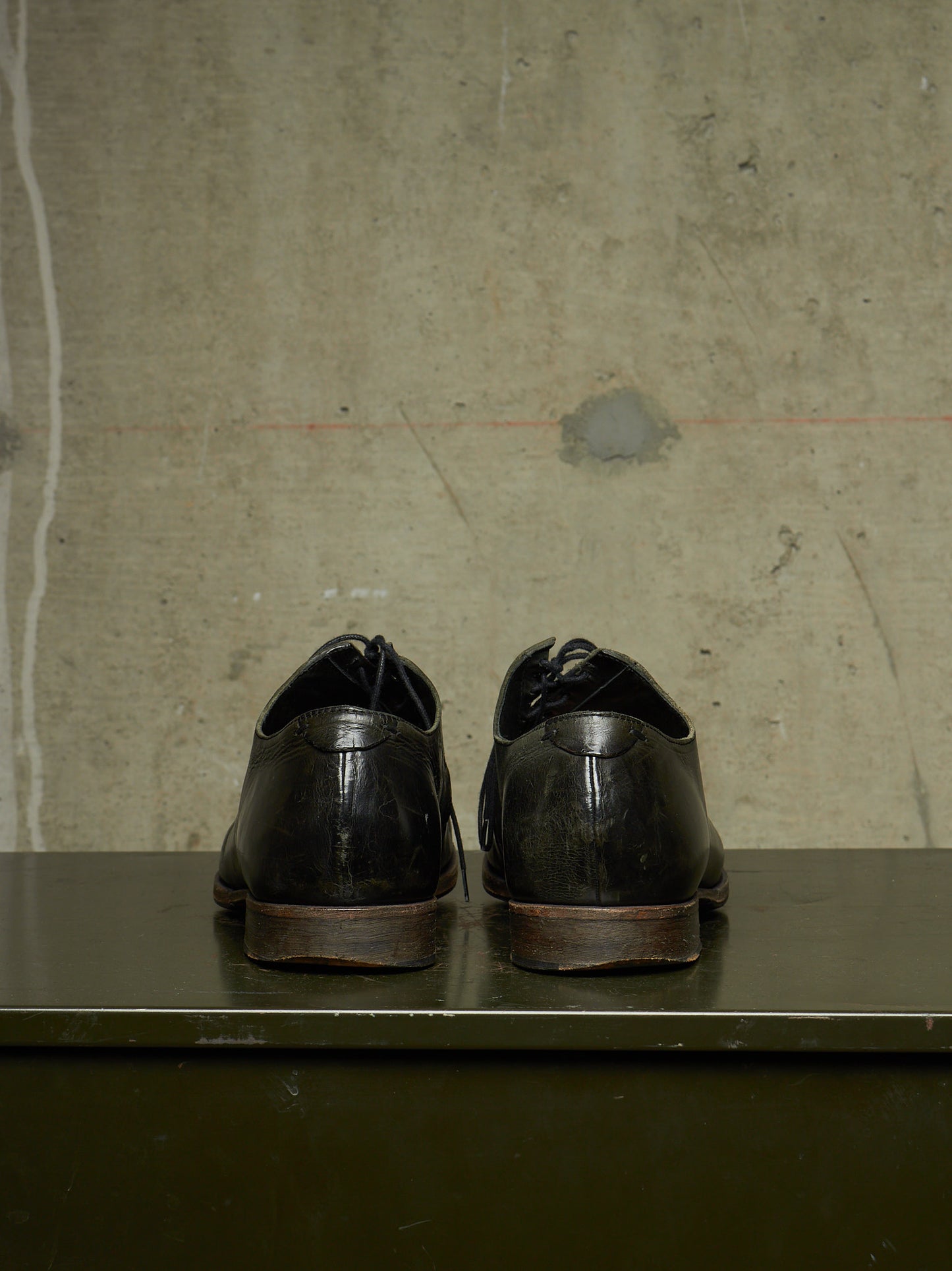 A pair of black leather dress shoes shown from the back, resting on a dark green surface against a textured concrete wall.