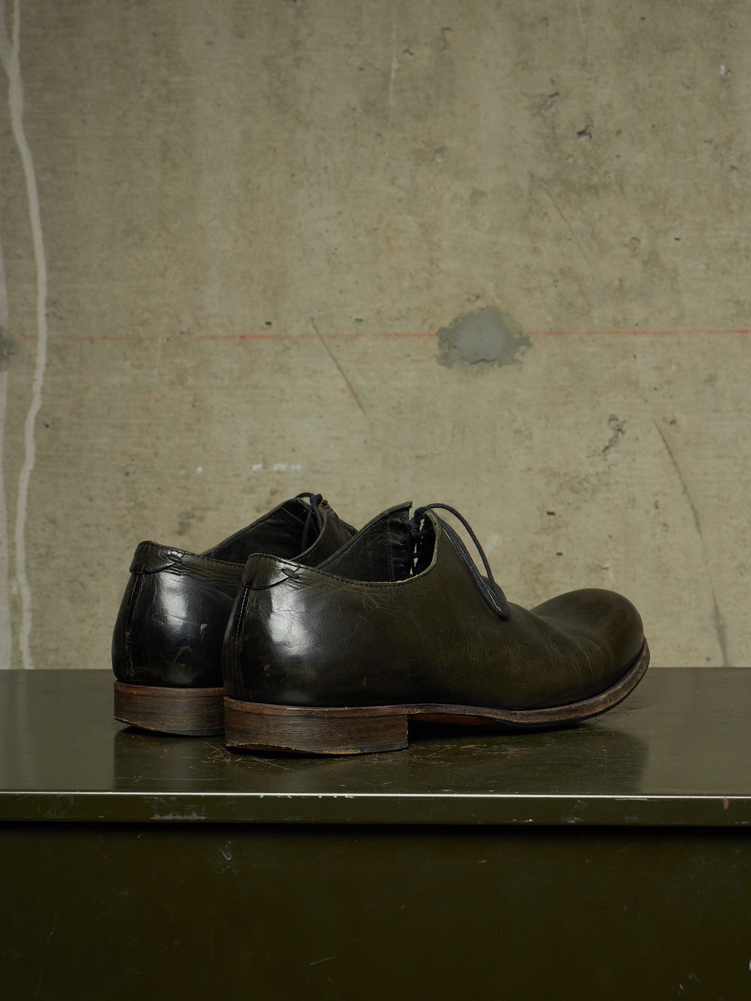 A pair of dark brown leather lace-up dress shoes with a low stacked heel, displayed on a dark green surface against a textured concrete wall.