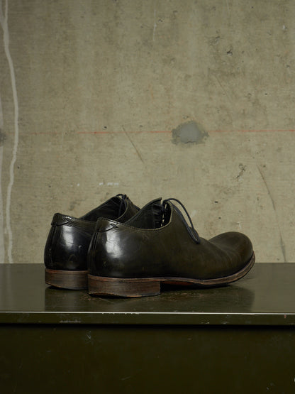 A pair of dark brown leather lace-up dress shoes with a low stacked heel, displayed on a dark green surface against a textured concrete wall.