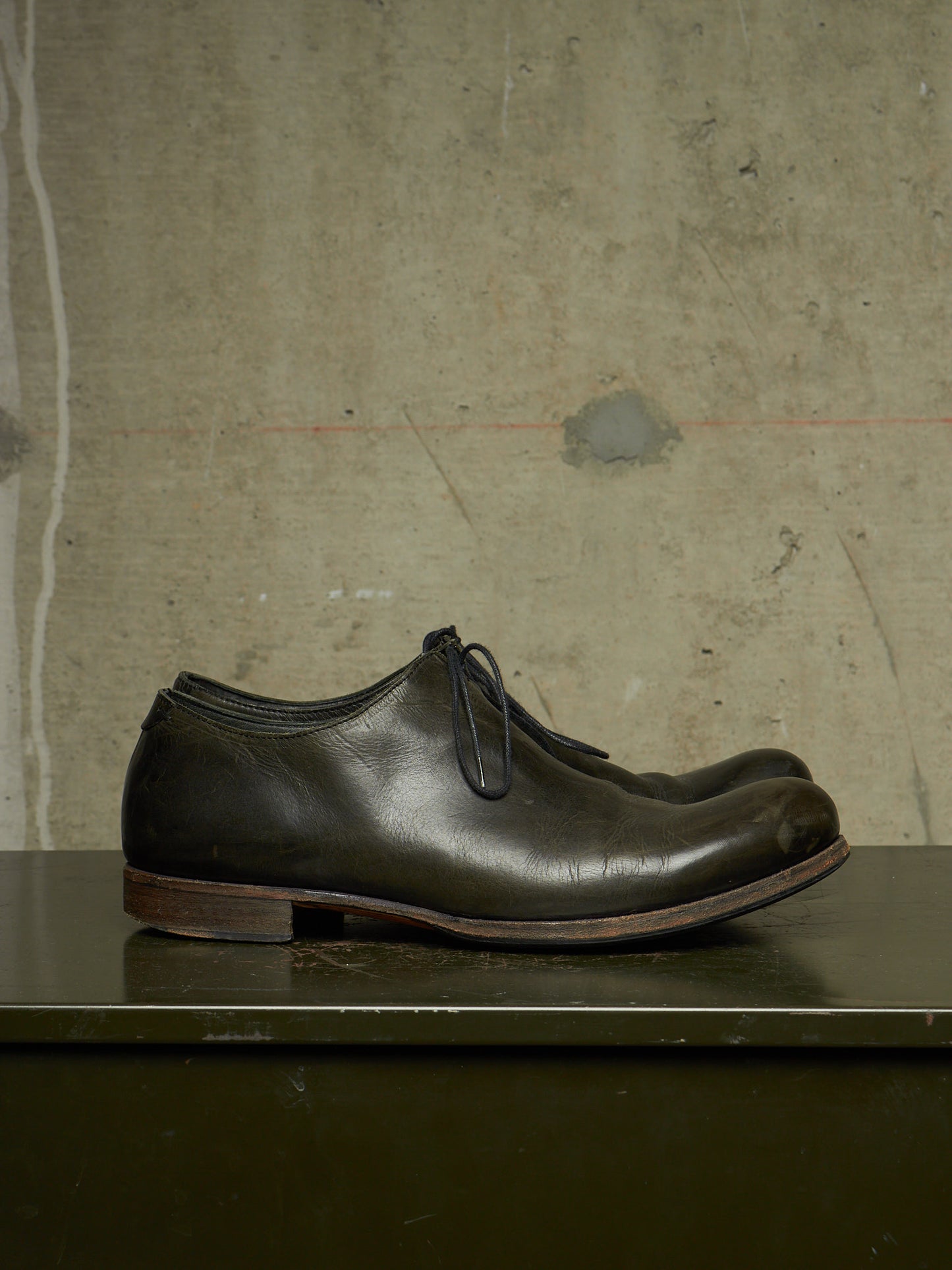A pair of dark brown leather lace-up dress shoes with a rounded toe, displayed on a dark surface against a textured concrete wall.