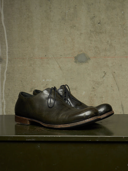A pair of dark brown leather lace-up dress shoes with a polished finish, displayed on a dark green surface against a textured concrete wall.