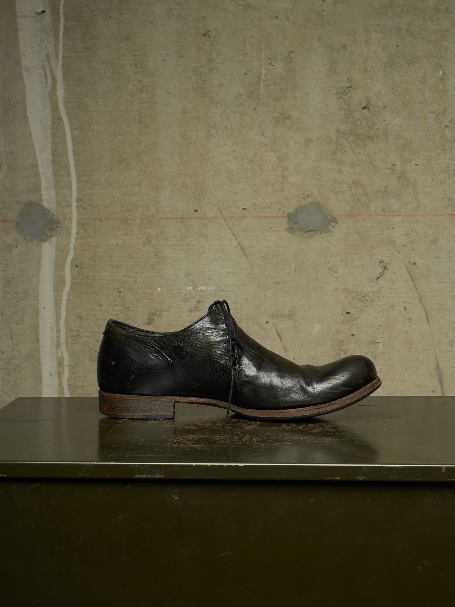 A single black leather lace-up dress shoe with a low heel, resting on a dark green surface against a textured concrete wall background.