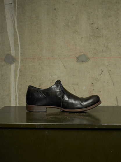 A single black leather lace-up dress shoe with a low heel, resting on a dark green surface against a textured concrete wall background.
