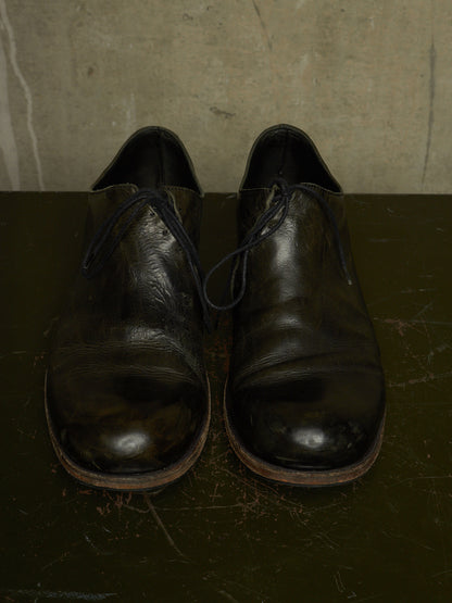 A pair of worn black leather cap-toe oxford shoes with dark laces, resting on a dark surface against a textured concrete wall.