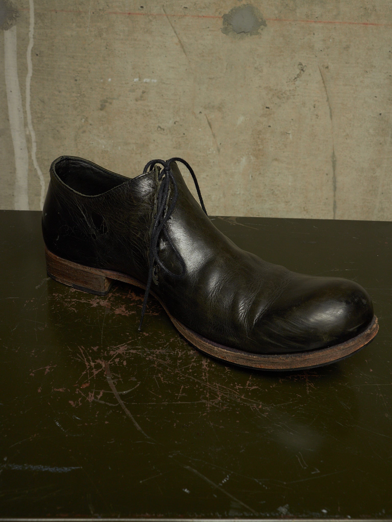 A single black leather lace-up ankle boot with a rounded toe and low stacked heel, resting on a dark scratched surface against a textured concrete wall.