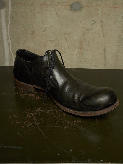 A single black leather lace-up ankle boot with a rounded toe and low stacked heel, resting on a dark scratched surface against a textured concrete wall.