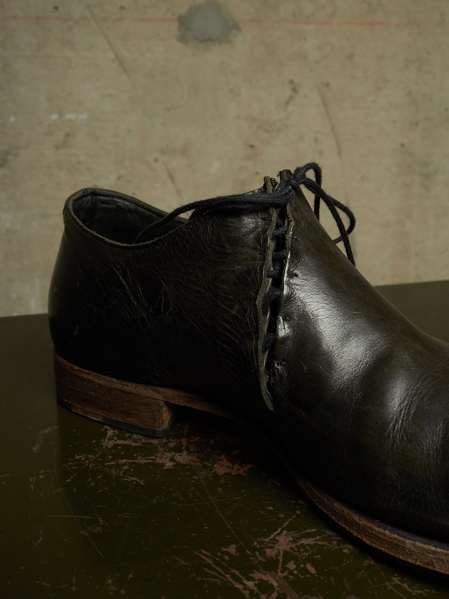 A single dark brown leather lace-up shoe photographed on a dark surface against a textured beige wall.