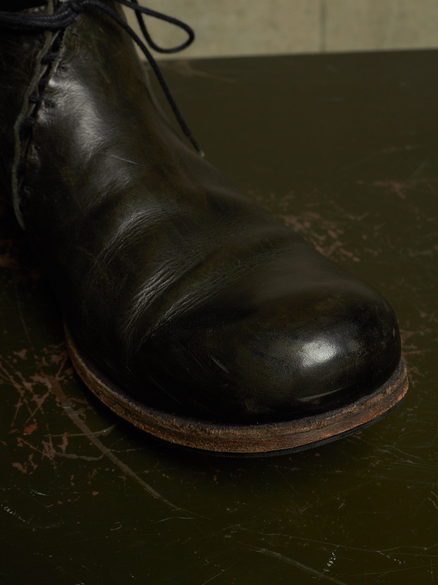 A close-up shot of a dark brown leather lace-up boot resting on a dark, scratched surface.