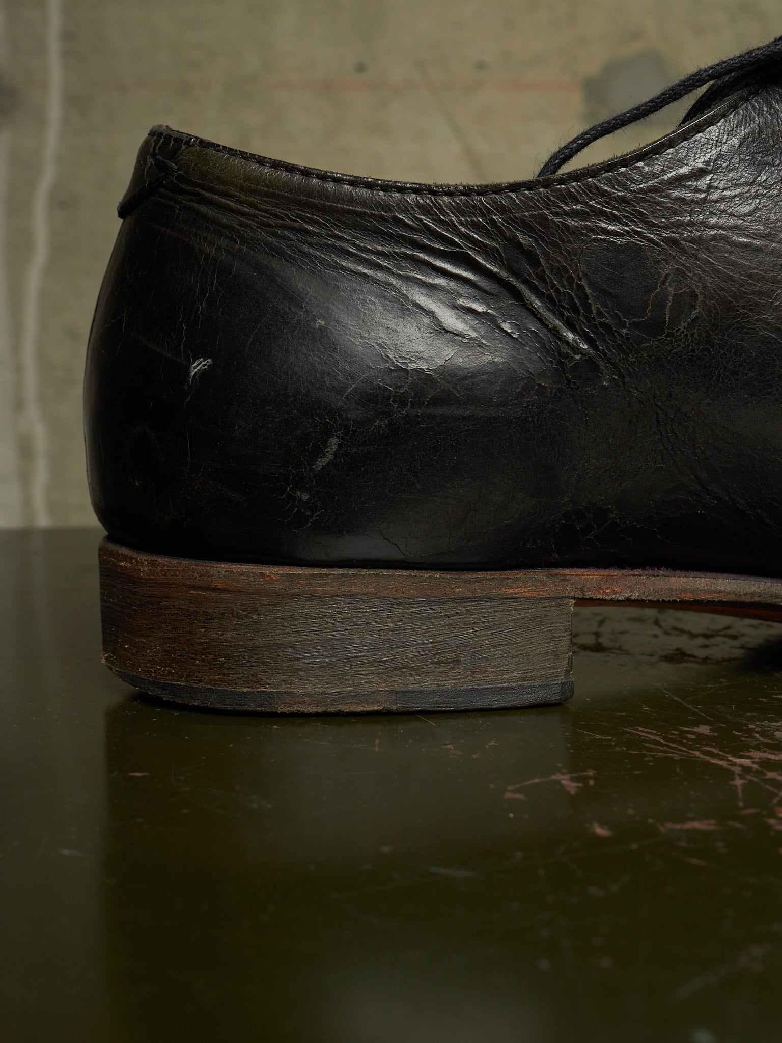 A close-up shot of the heel and side of a worn black leather dress shoe resting on a dark reflective surface.