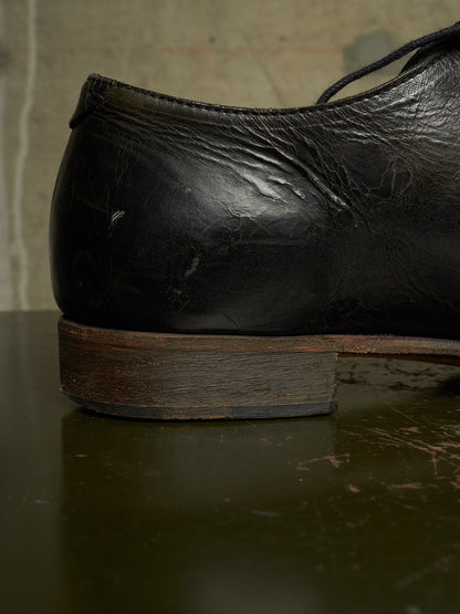 A close-up shot of the heel and side of a worn black leather dress shoe resting on a dark reflective surface.