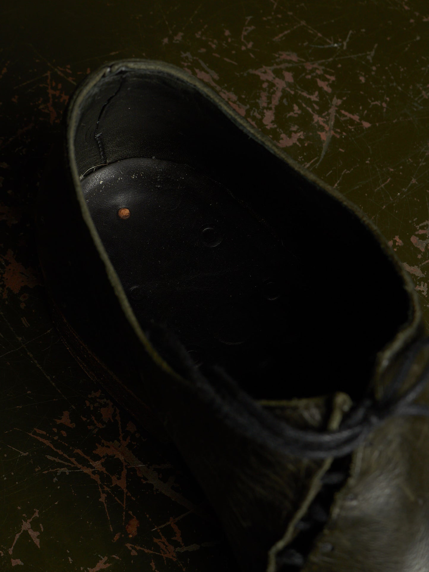 A close-up, high-angle view of the dark interior opening of a black lace-up shoe, set against a dark, distressed olive green background.