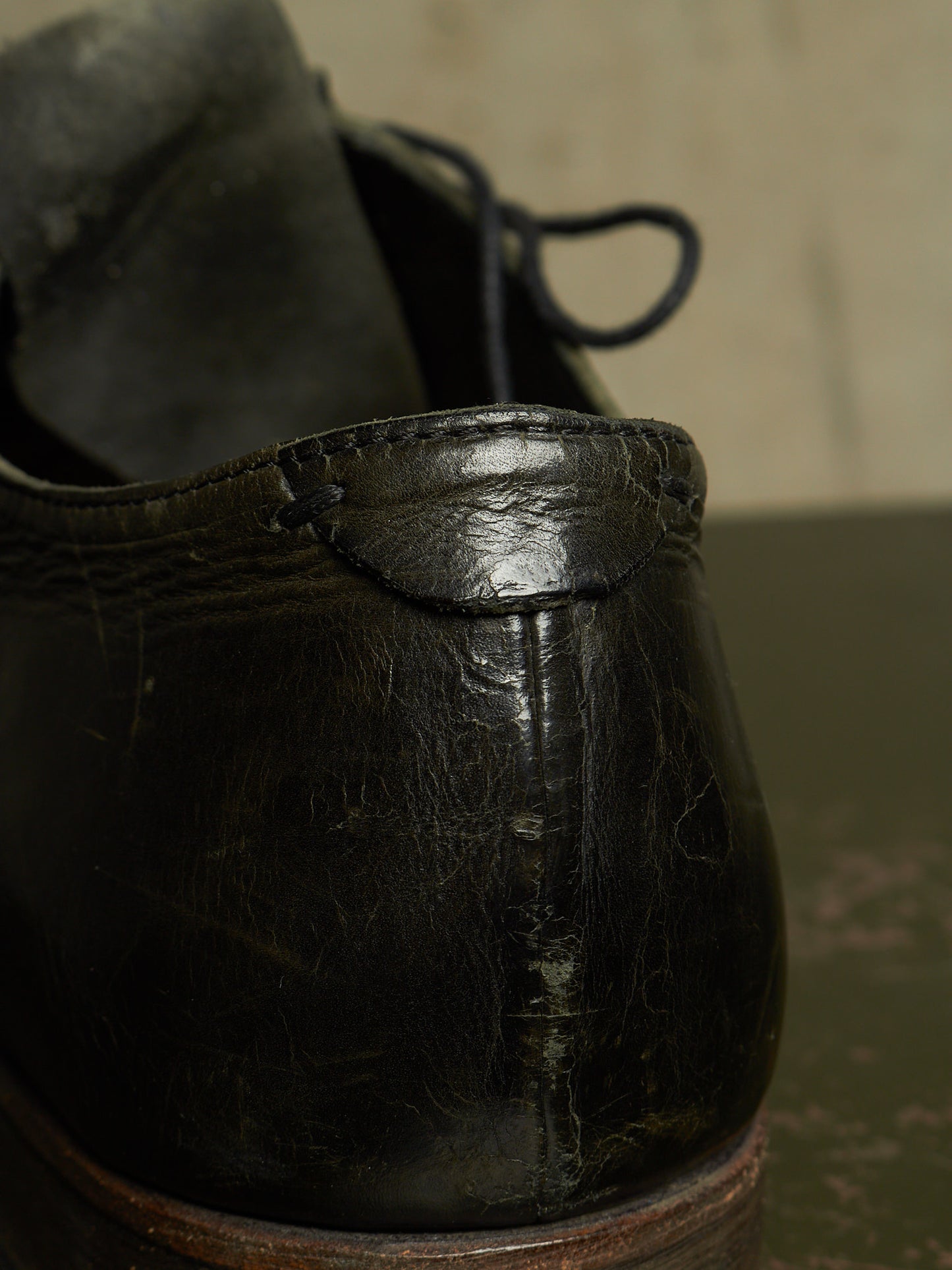 A close-up shot of a worn black leather lace-up shoe, showing the heel and side profile with visible scuffs and creases, set against a dark background.