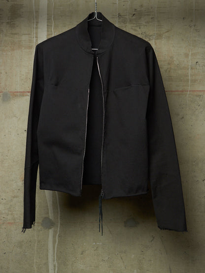 A black zip-up jacket with a stand collar and raw, frayed hems, hanging against a textured concrete wall.