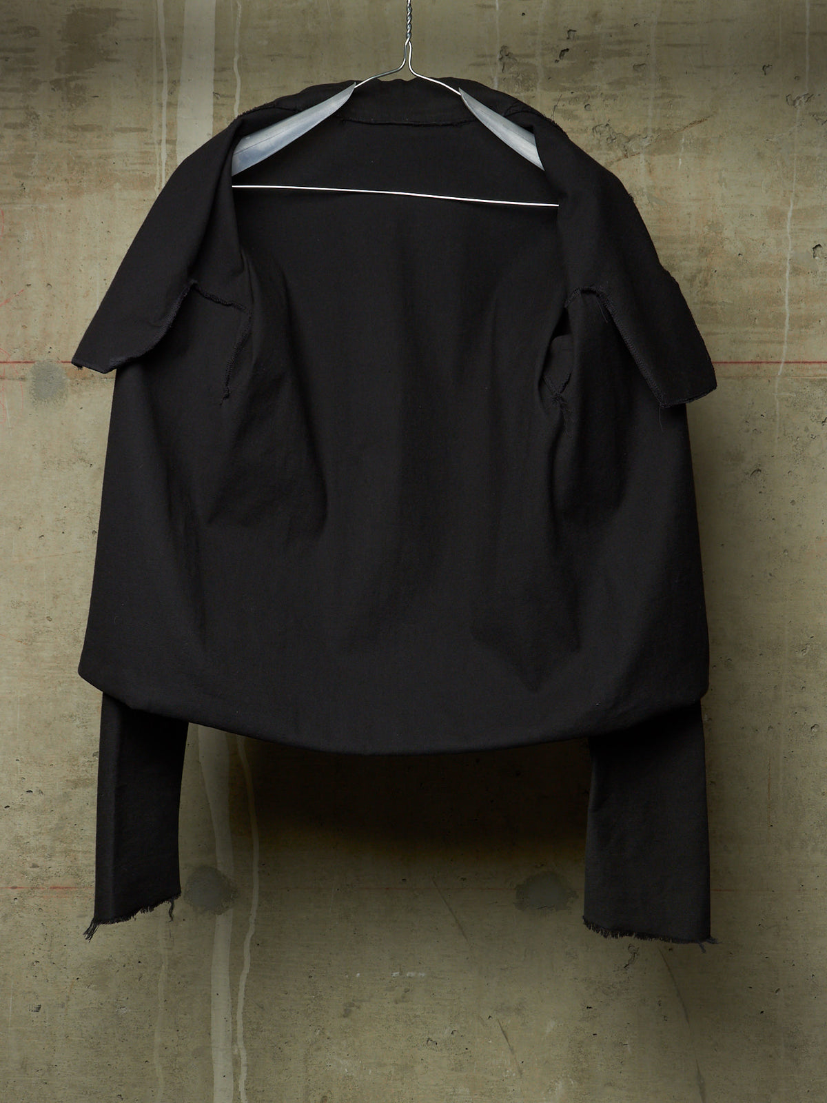 A black oversized cropped jacket with wide lapels and long draped sleeves, hanging on a metal hanger against a textured concrete wall.