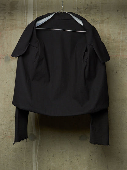 A black oversized cropped jacket with wide lapels and long draped sleeves, hanging on a metal hanger against a textured concrete wall.