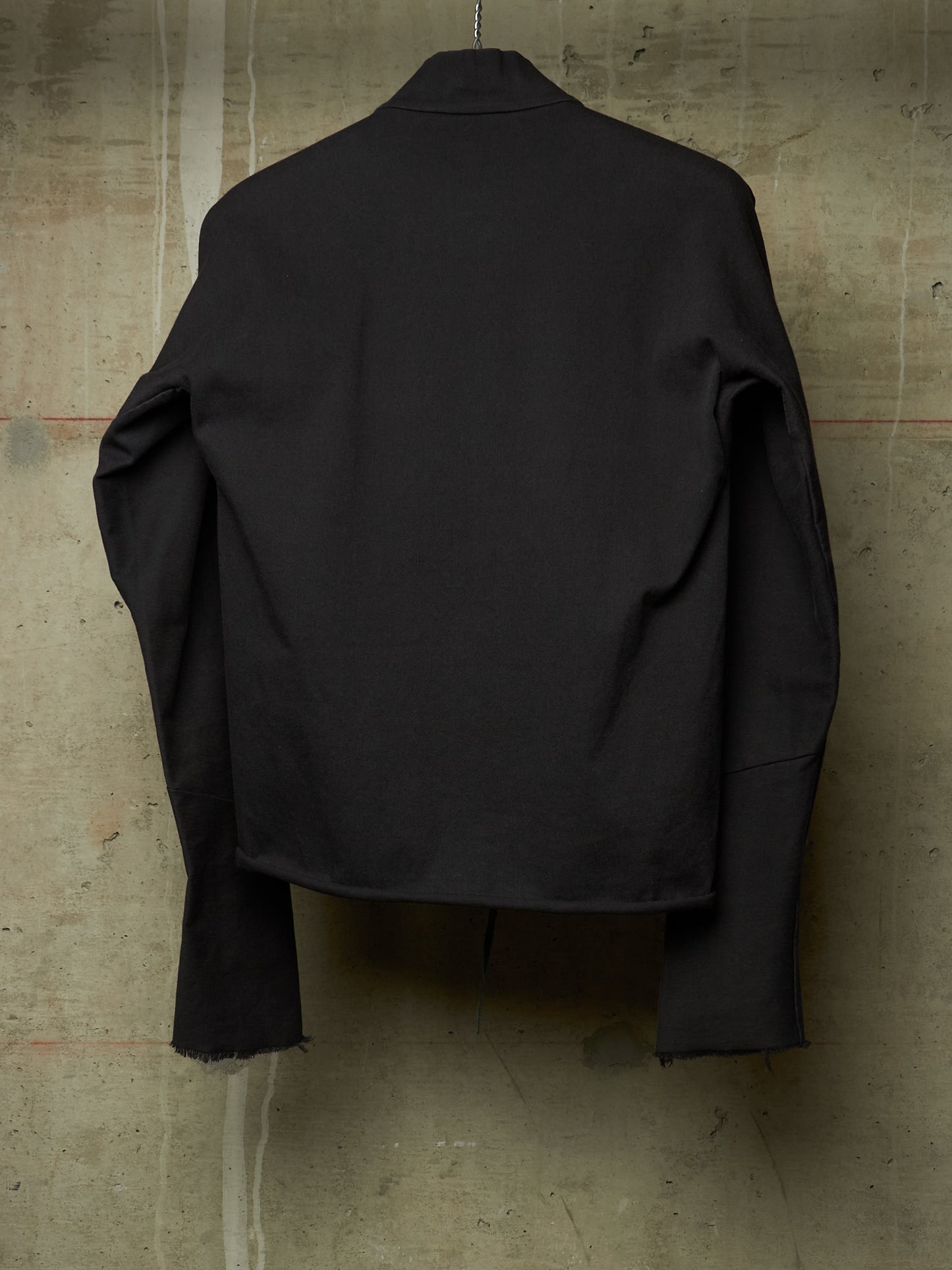 A black long-sleeve cropped jacket or overshirt hanging against a textured concrete wall, shown from the back.