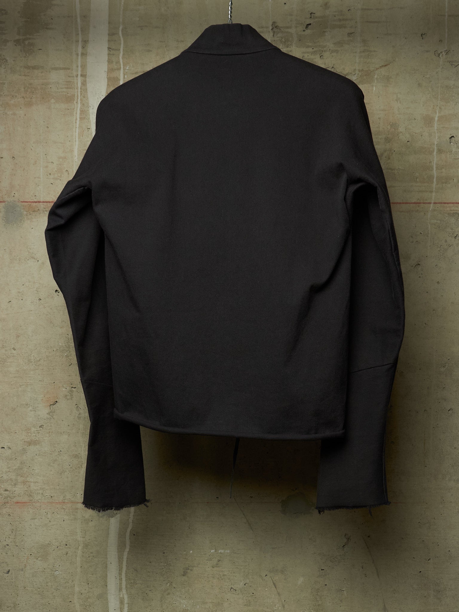 A black long-sleeve cropped jacket or overshirt hanging against a textured concrete wall, shown from the back.