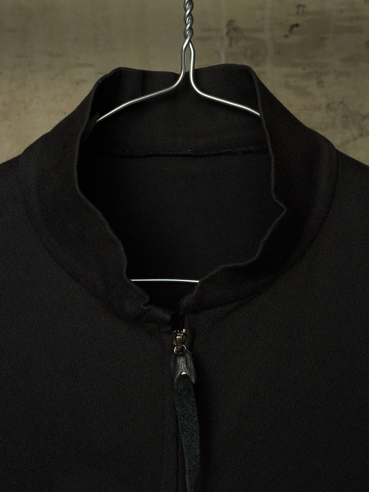 A close-up of a black knit zip-up sweater hanging on a metal hanger against a textured, warm-toned background.