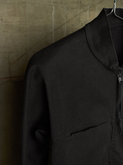 A close-up of a black zip-up jacket with a high collar, hanging on a metal hanger against a textured concrete wall.