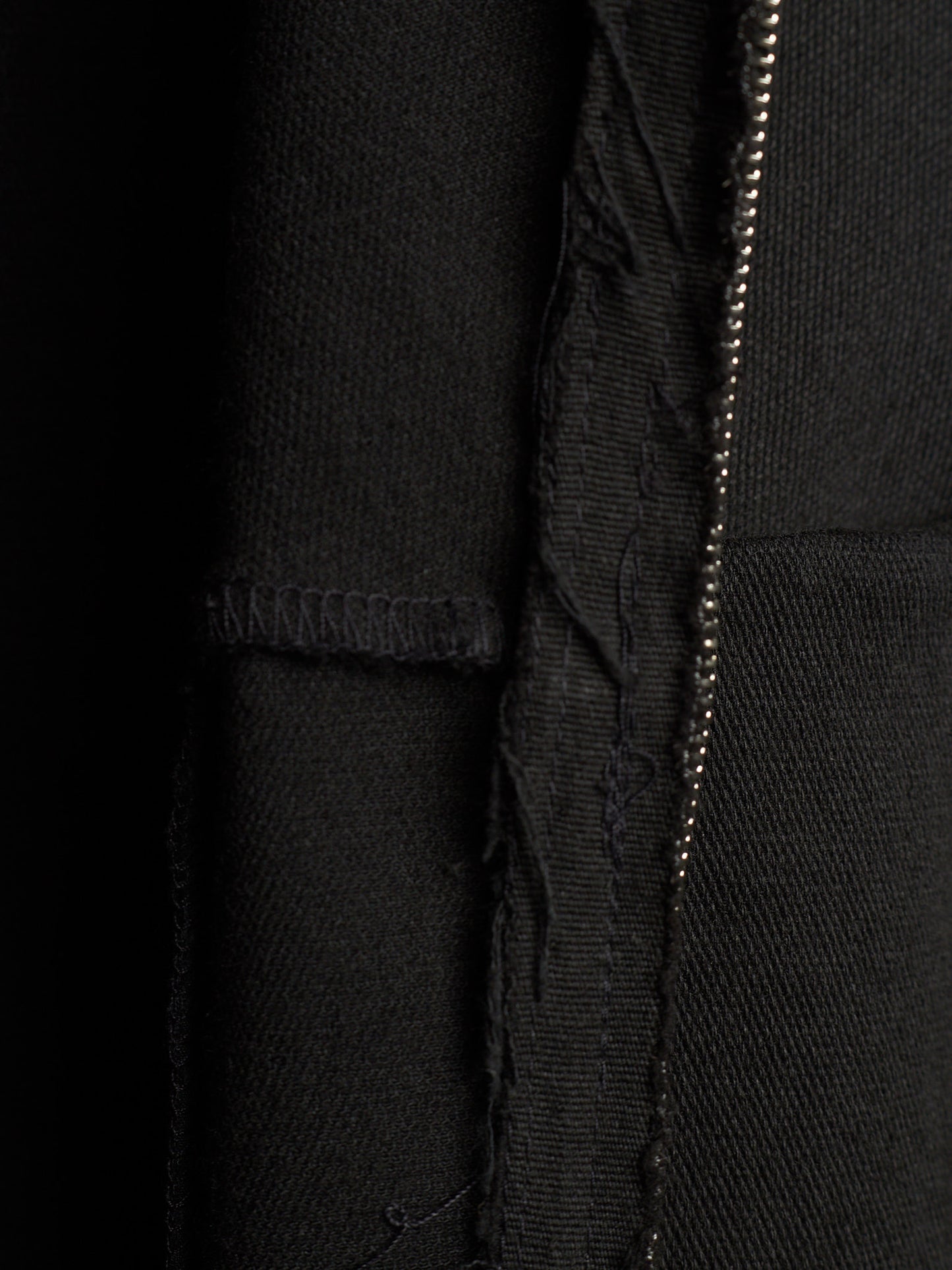 Close-up detail of a black textured fabric garment showing a vertical seam, raw frayed edge, and a metallic zipper running down the center.