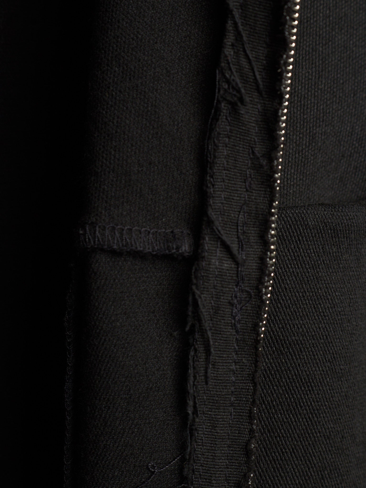 Close-up detail of a black textured fabric garment showing a vertical seam, raw frayed edge, and a metallic zipper running down the center.