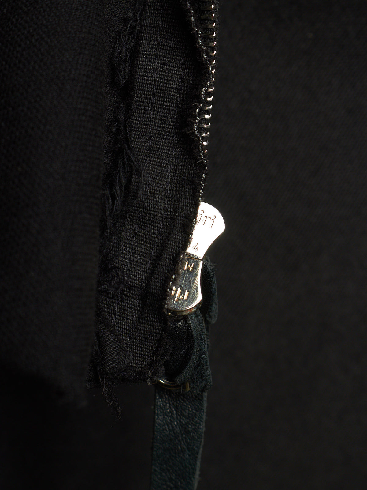 Close-up of a black fabric garment with a distressed, frayed edge and a silver metal zipper pull, set against a dark black background.