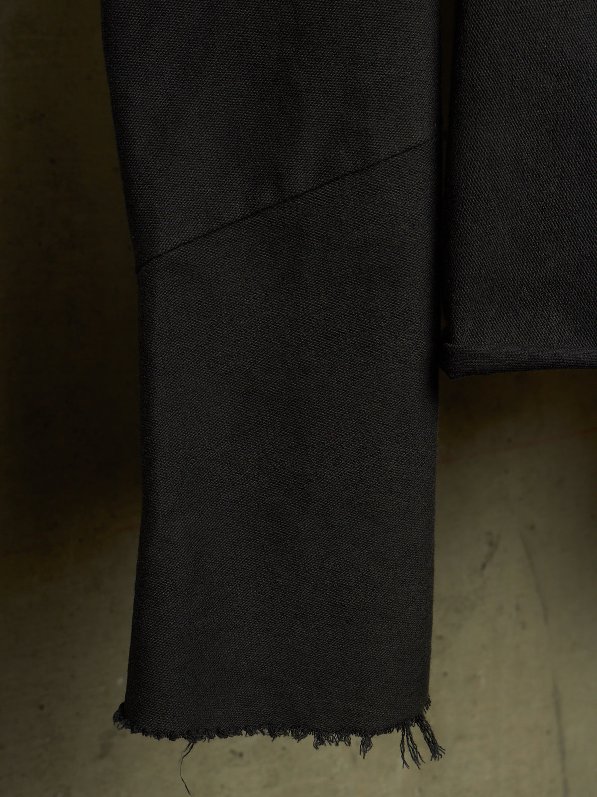 Close-up of a black fabric garment sleeve or drape with a raw, frayed hem, shown against a neutral concrete floor.