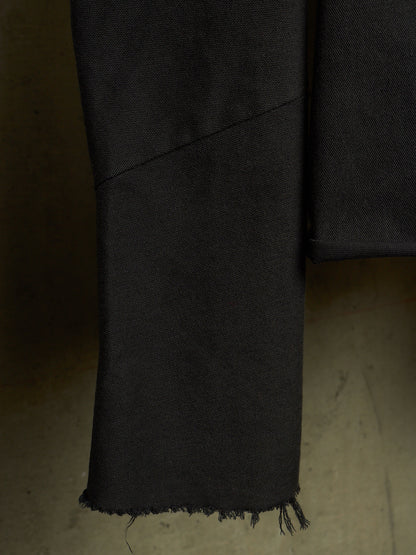 Close-up of a black fabric garment sleeve or drape with a raw, frayed hem, shown against a neutral concrete floor.