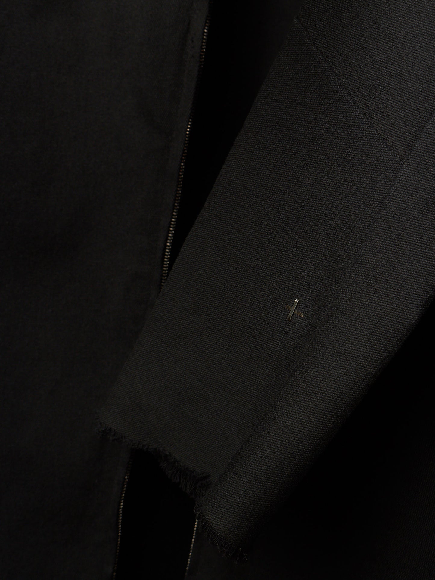 Close-up detail of a black textured jacket or coat showing a sleeve, raw frayed hem, and a small metallic logo pin.