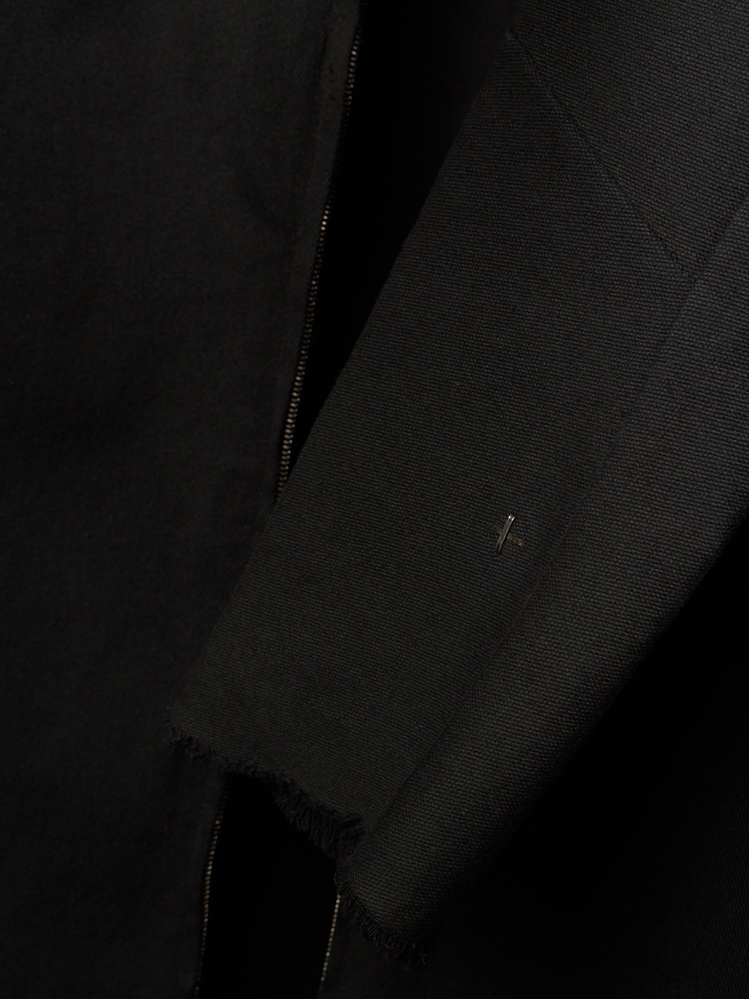 Close-up detail of a black textured jacket or coat showing a sleeve, raw frayed hem, and a small metallic logo pin.