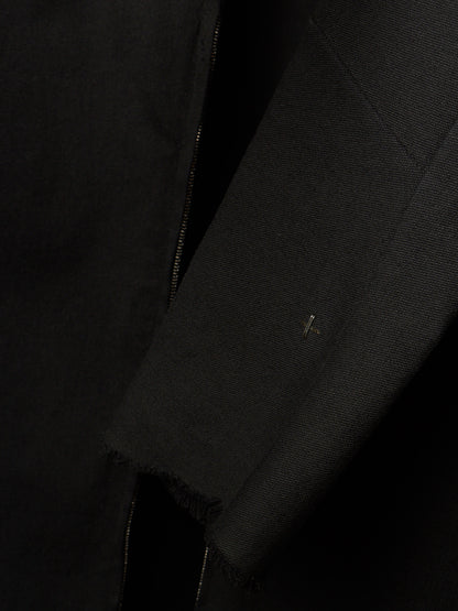 Close-up detail of a black textured jacket or coat showing a sleeve, raw frayed hem, and a small metallic logo pin.