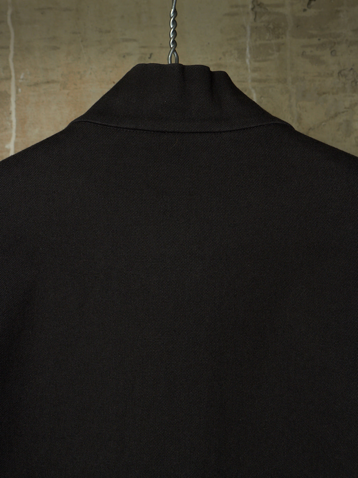 Close-up of the back of a black knit garment hanging on a metal hanger against a textured beige wall.