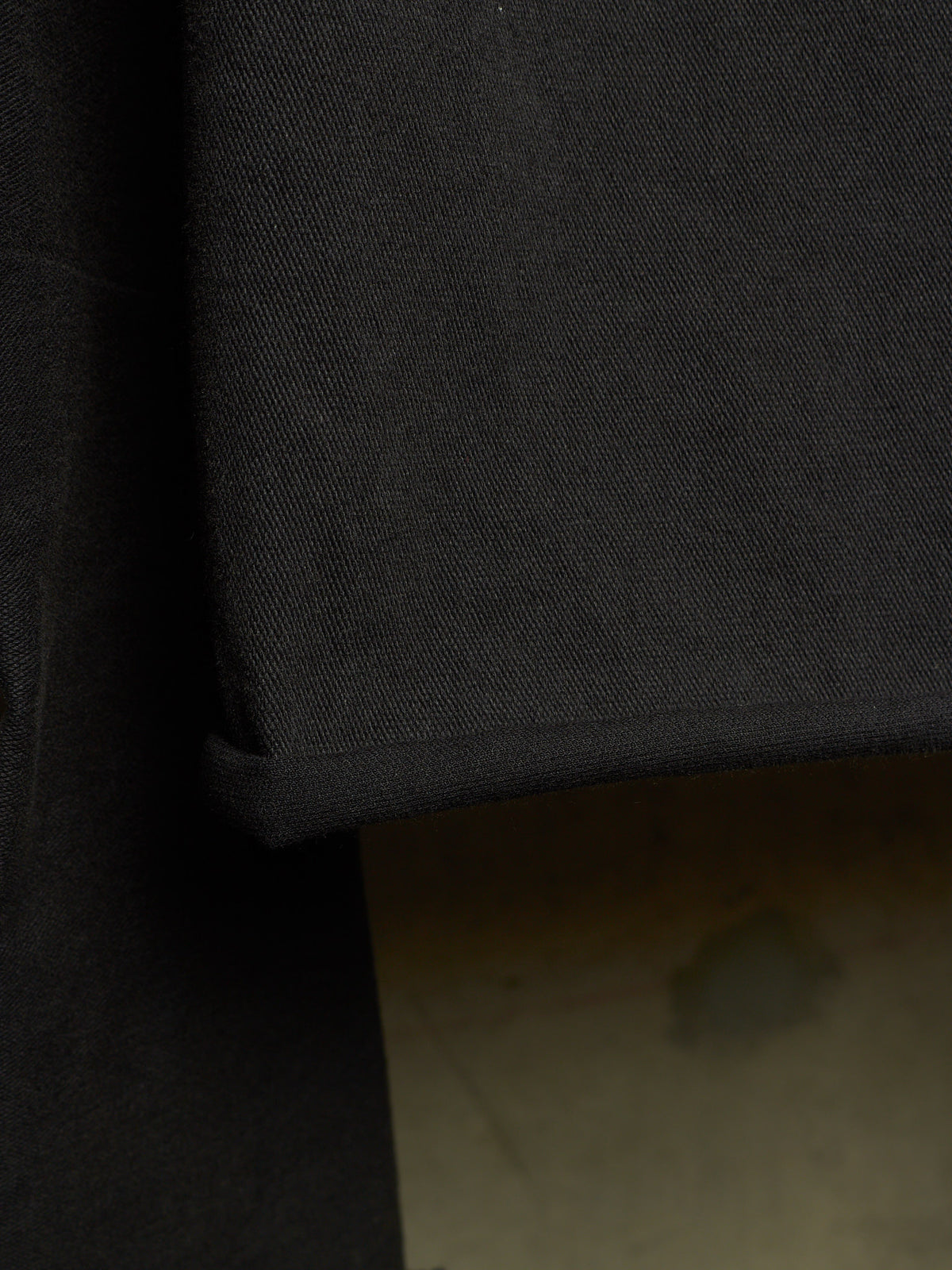 Close-up detail of a black fabric garment, showing the texture of the material and a folded cuff, with a neutral floor visible in the background.
