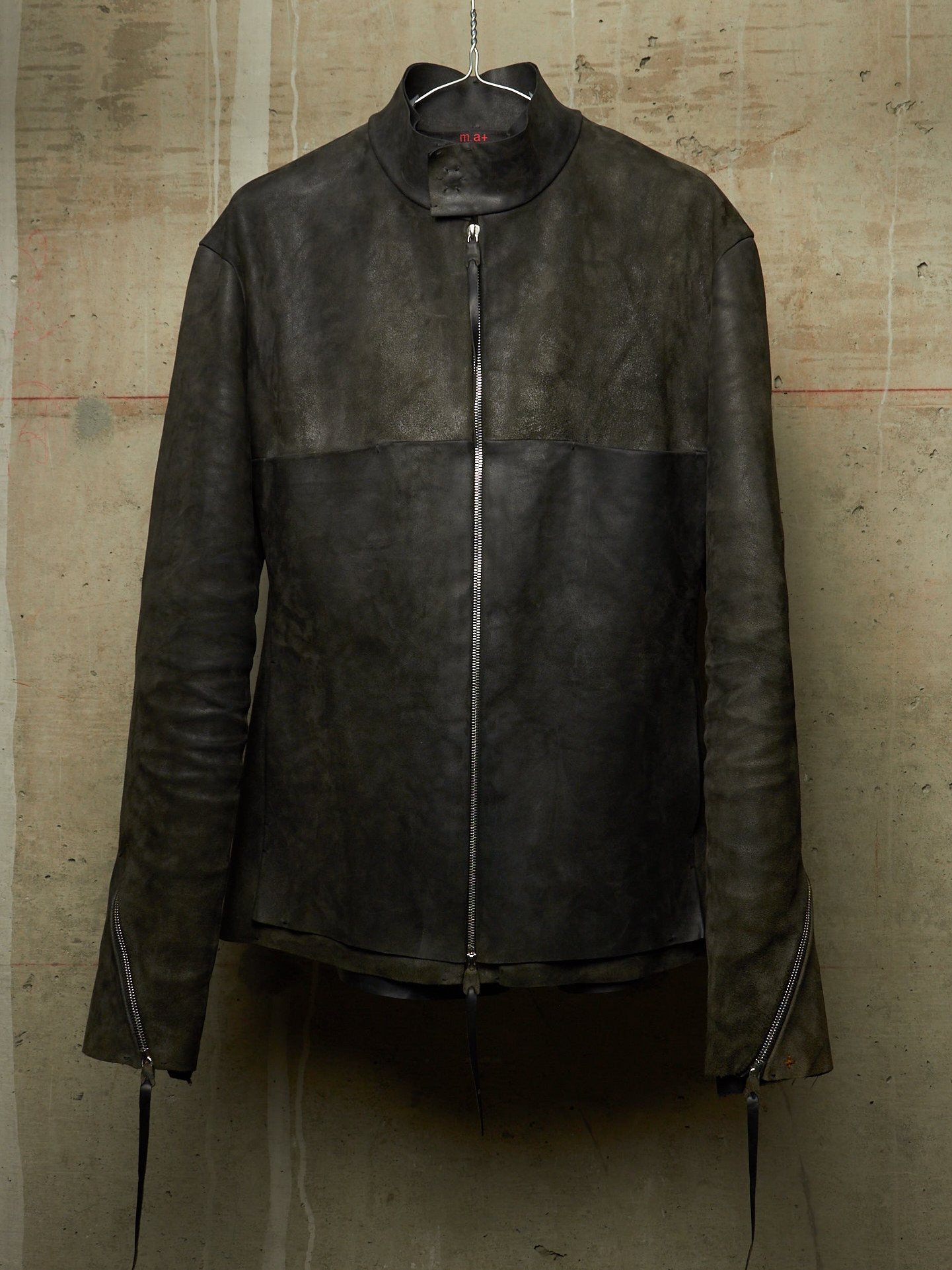 A dark brown leather jacket with a high mock neck collar and front zipper, hanging on a wire hanger against a textured concrete wall.