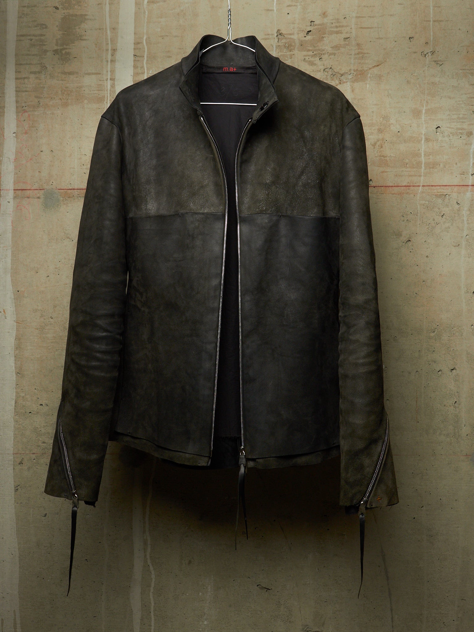A dark olive green leather jacket with a split-color panel design, hanging on a wire hanger against a textured concrete wall.