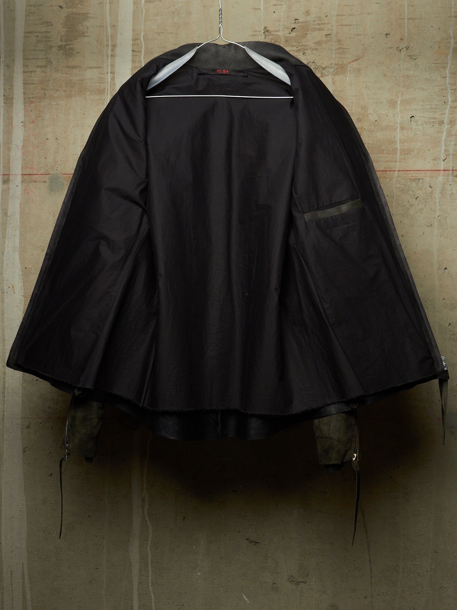 A black hooded raincoat or cape-style outerwear garment hanging on a hanger against a textured concrete wall. The hood is up, and a side pocket is visible on the right side of the garment.