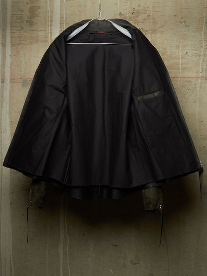 A black hooded raincoat or cape-style outerwear garment hanging on a hanger against a textured concrete wall. The hood is up, and a side pocket is visible on the right side of the garment.