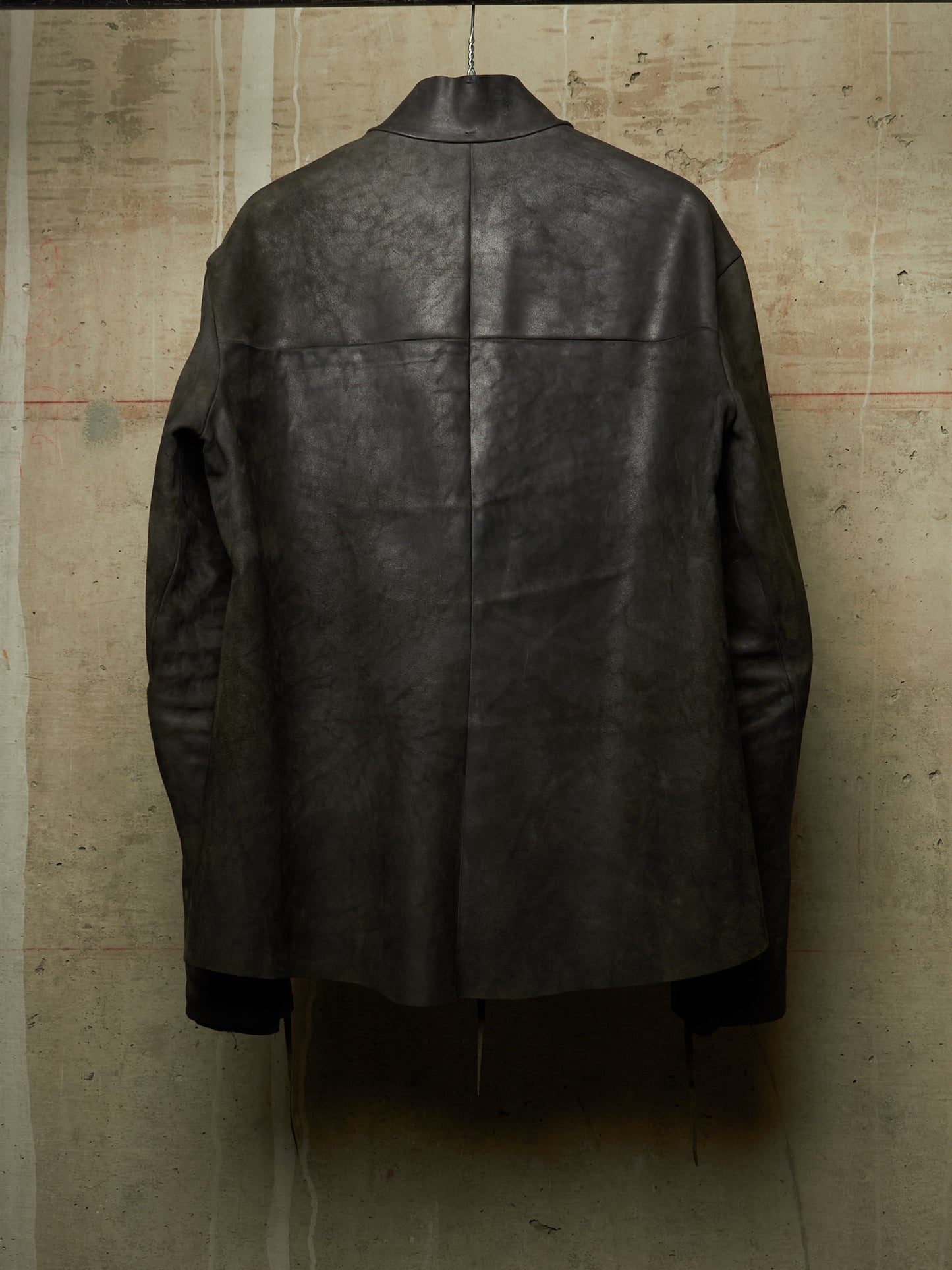 A dark charcoal gray distressed leather jacket shown from the back, hanging against a textured concrete wall.