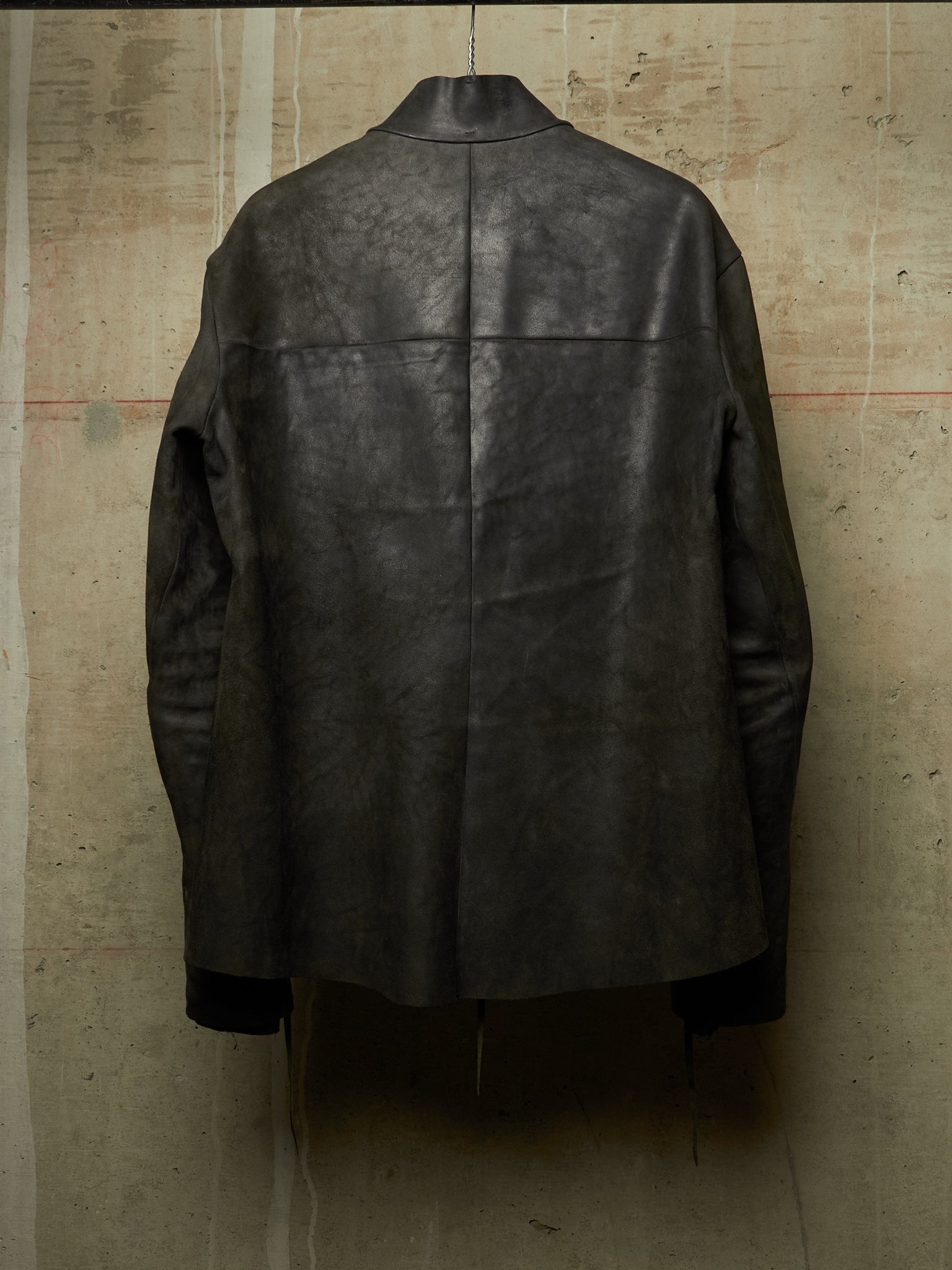 A dark charcoal gray distressed leather jacket shown from the back, hanging against a textured concrete wall.