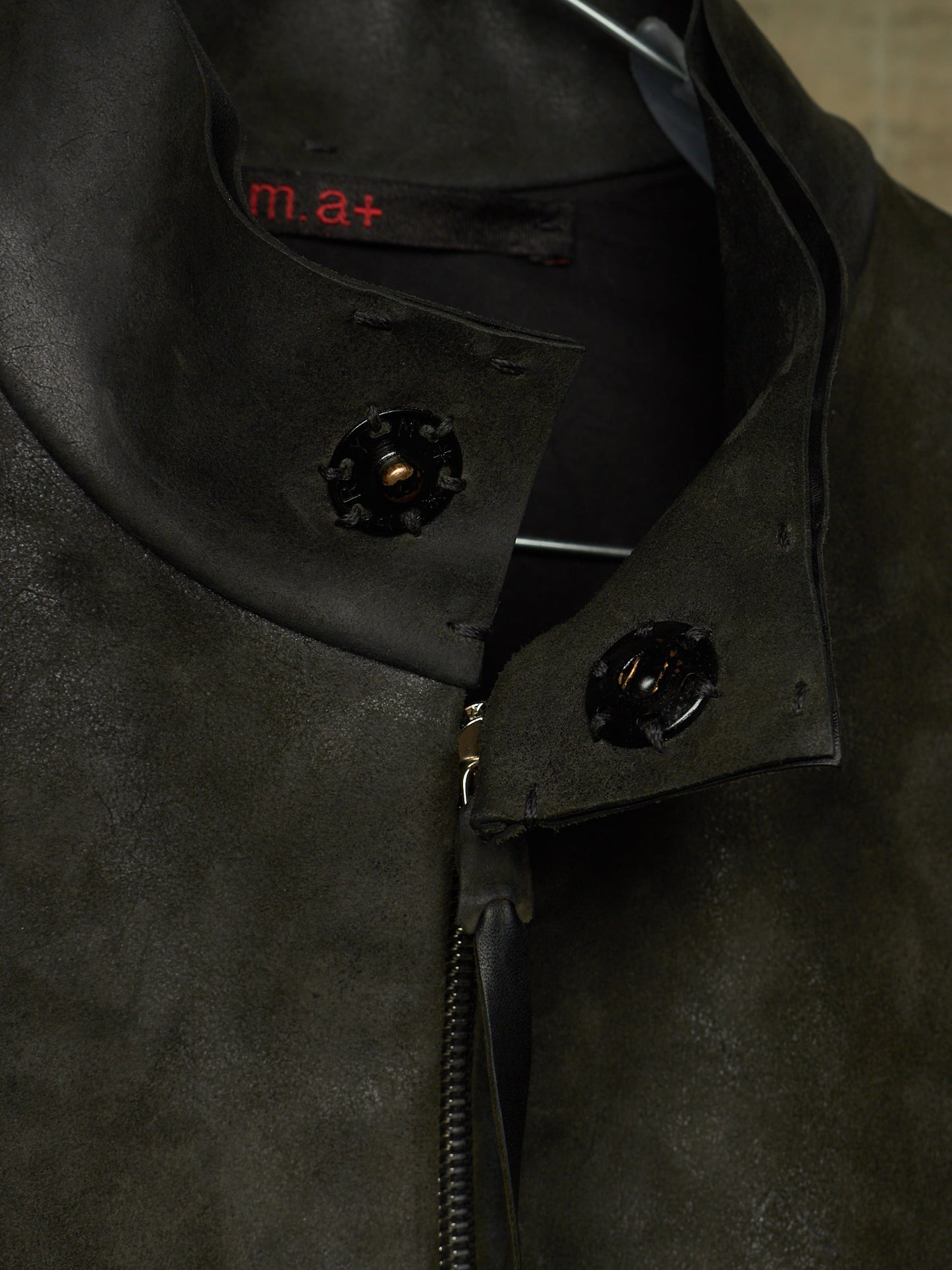 Close-up of a dark olive green suede jacket with a high collar, black decorative snap buttons, and a black zipper. The brand name 'm.