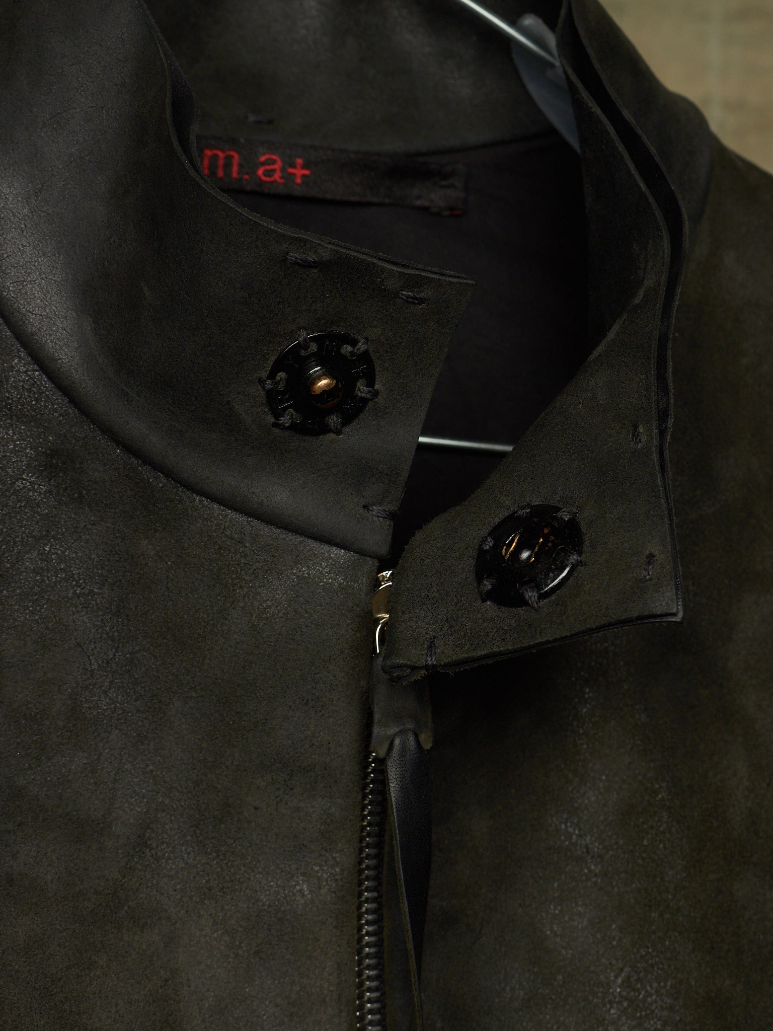 Close-up of a dark olive green suede jacket with a high collar, black decorative snap buttons, and a black zipper. The brand name 'm.