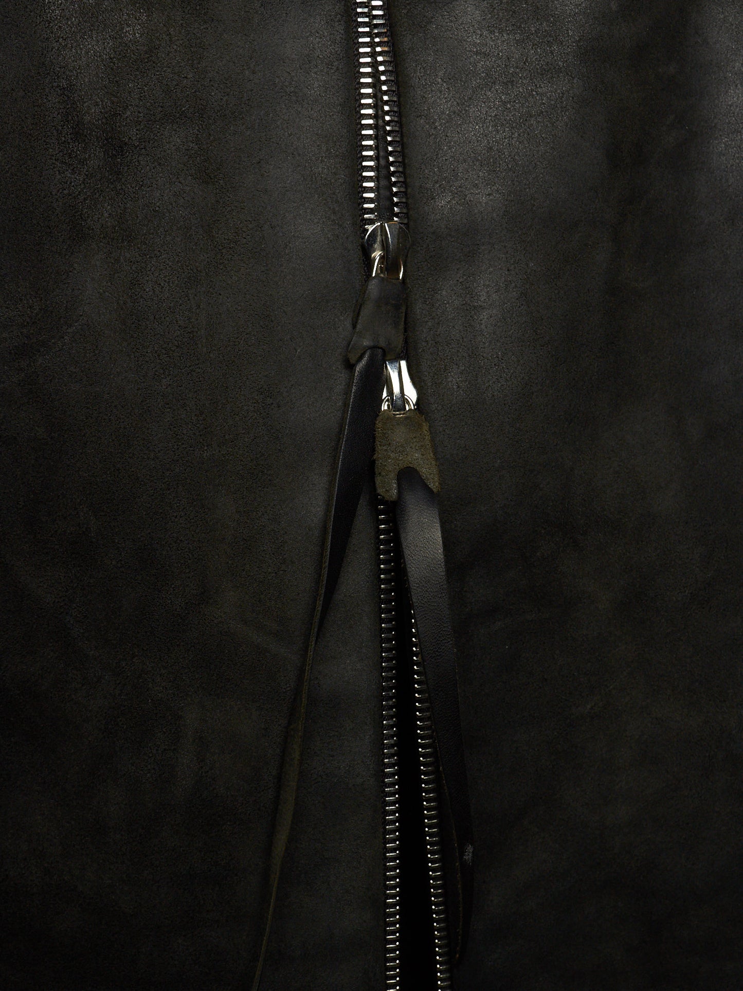 Close-up of a black zipper with a black leather pull tab on a dark textured fabric background.