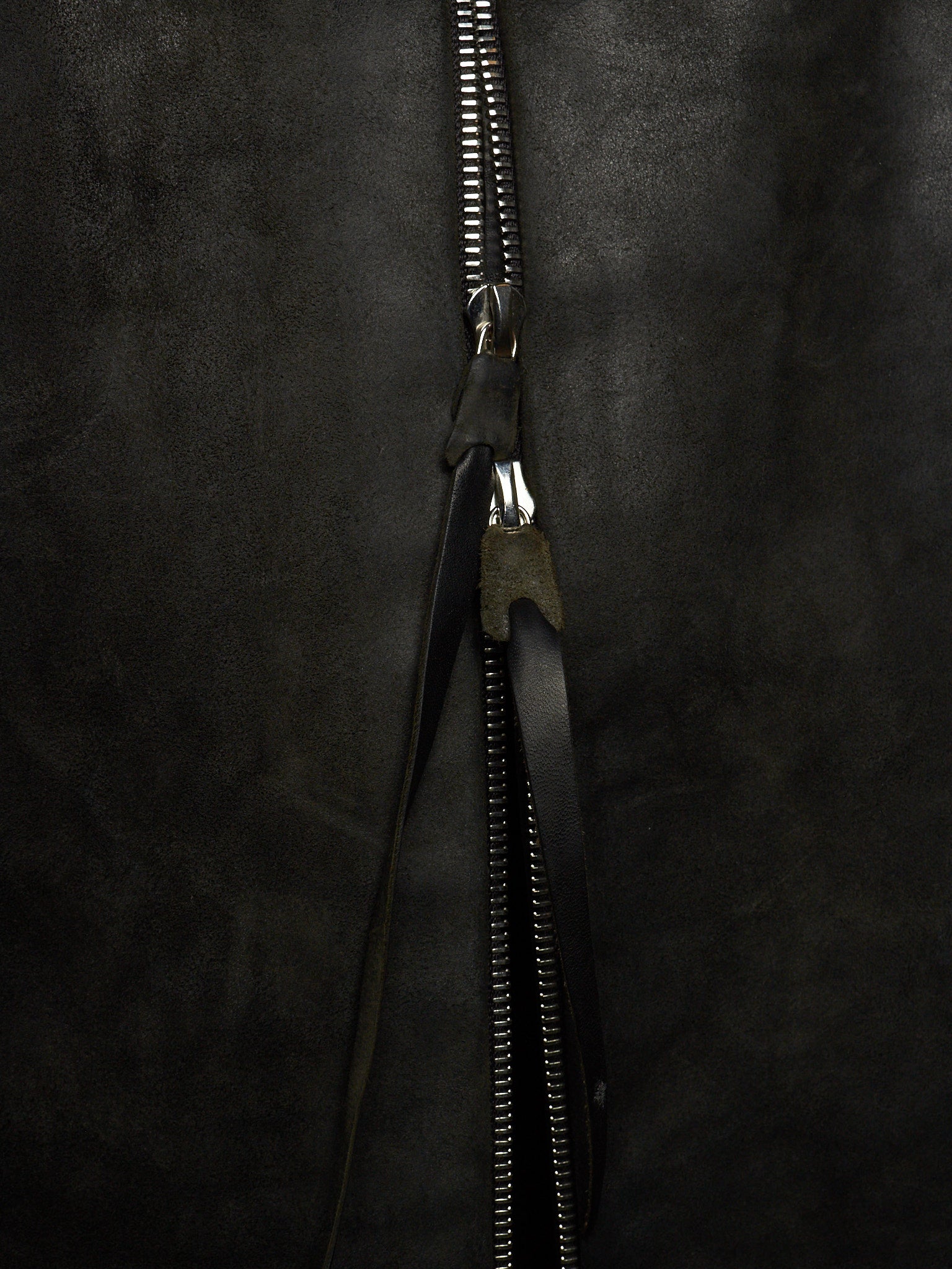 Close-up of a black zipper with a black leather pull tab on a dark textured fabric background.