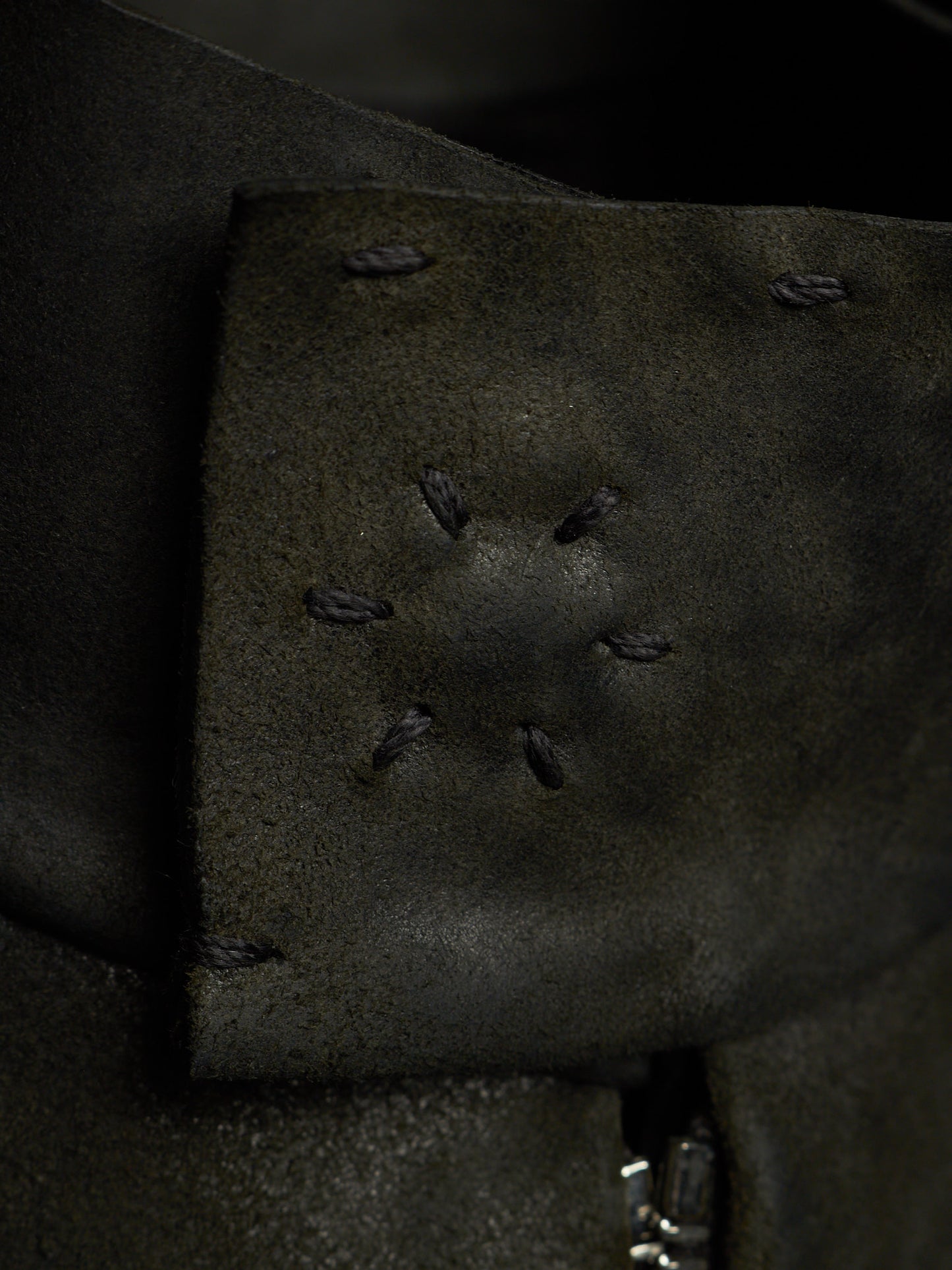 Close-up of a dark olive green leather strap or cuff with decorative whipstitching and a small silver metal clasp visible at the bottom edge.