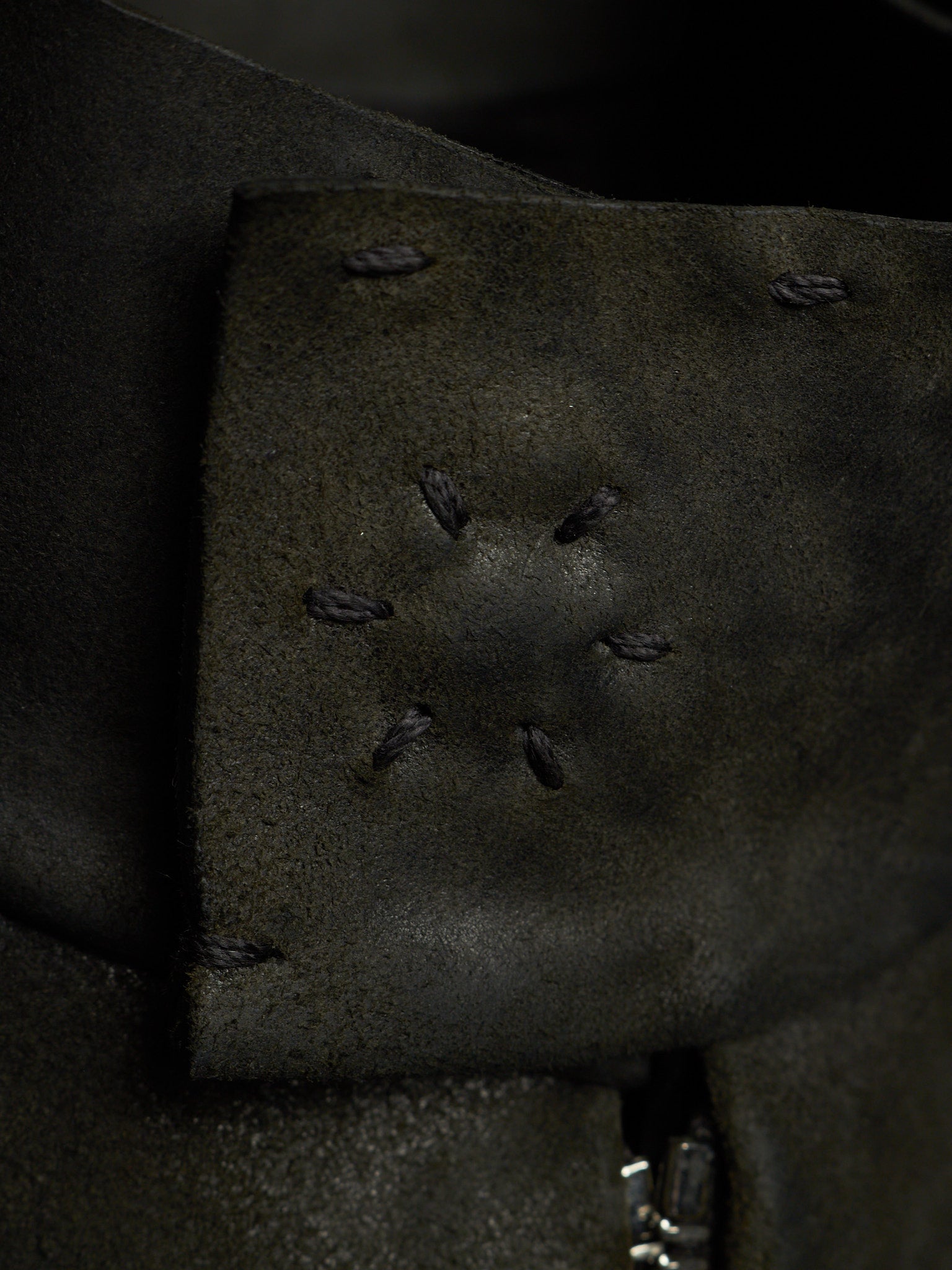Close-up of a dark olive green leather strap or cuff with decorative whipstitching and a small silver metal clasp visible at the bottom edge.