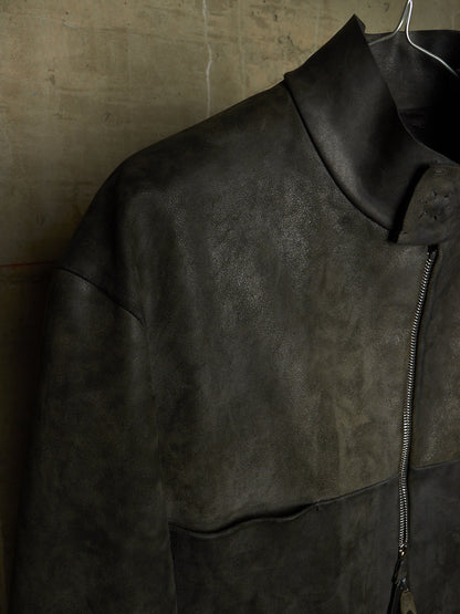 A dark charcoal gray leather jacket with a high collar and front zipper, hanging on a wire hanger against a textured concrete wall.