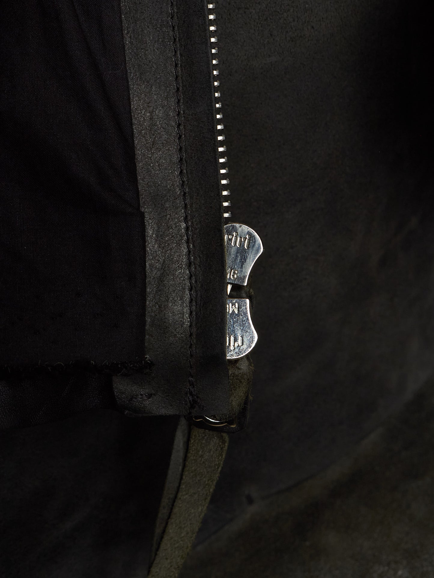 Close-up of a black leather jacket with a silver metal zipper and a decorative silver zipper pull featuring the brand name 'MUT'.