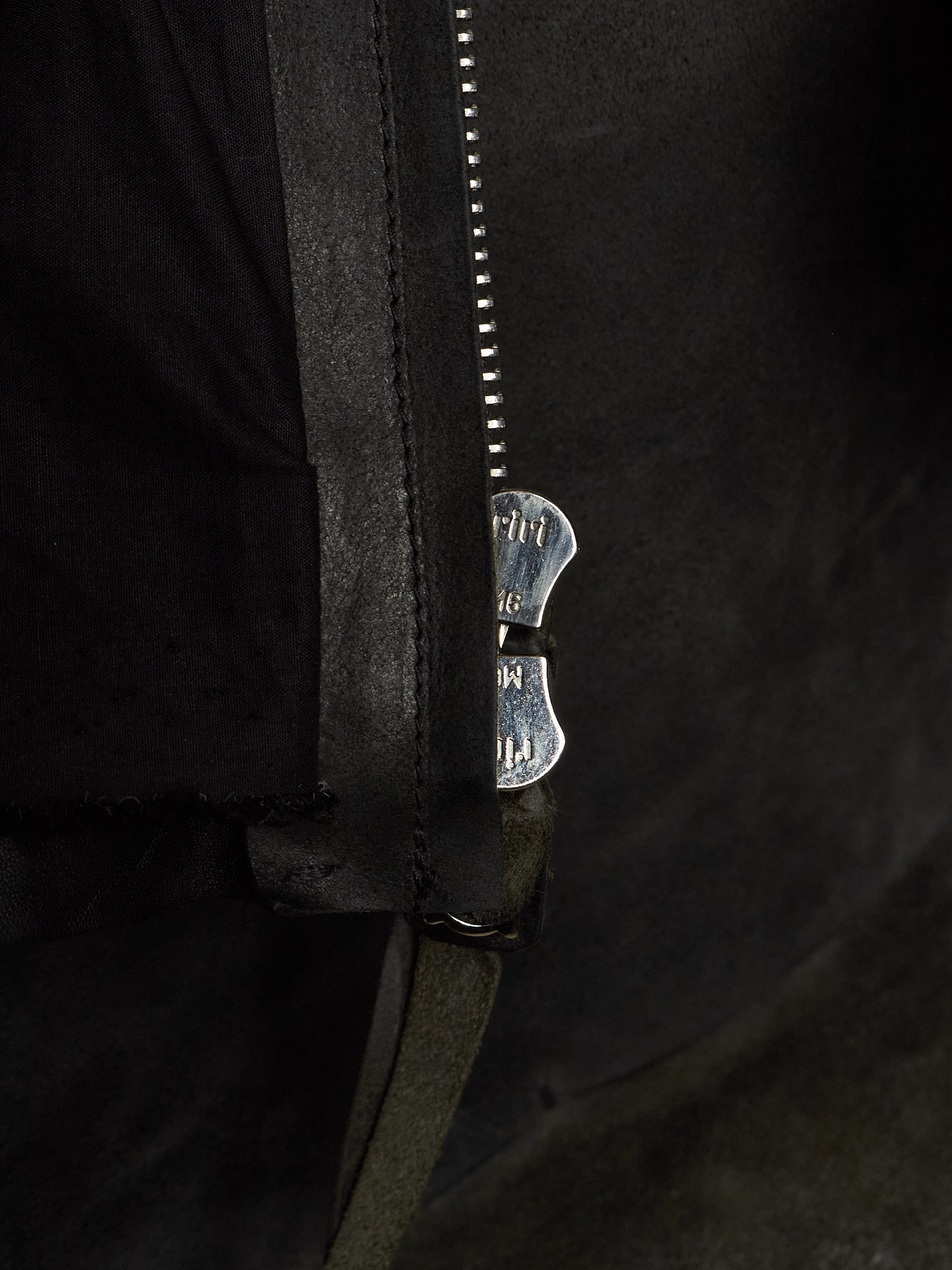 Close-up of a black leather jacket with a silver metal zipper and a decorative silver zipper pull featuring the brand name 'MUT'.
