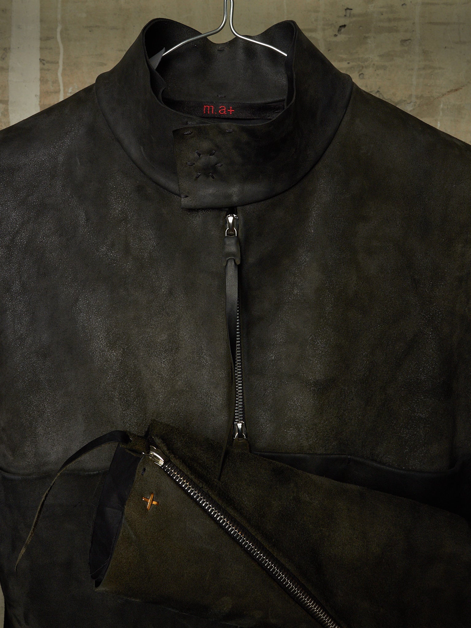 A dark brown suede half-zip jacket with a high collar, hanging on a metal hanger against a textured concrete wall. A matching dark brown suede zippered pouch is draped in the foreground.