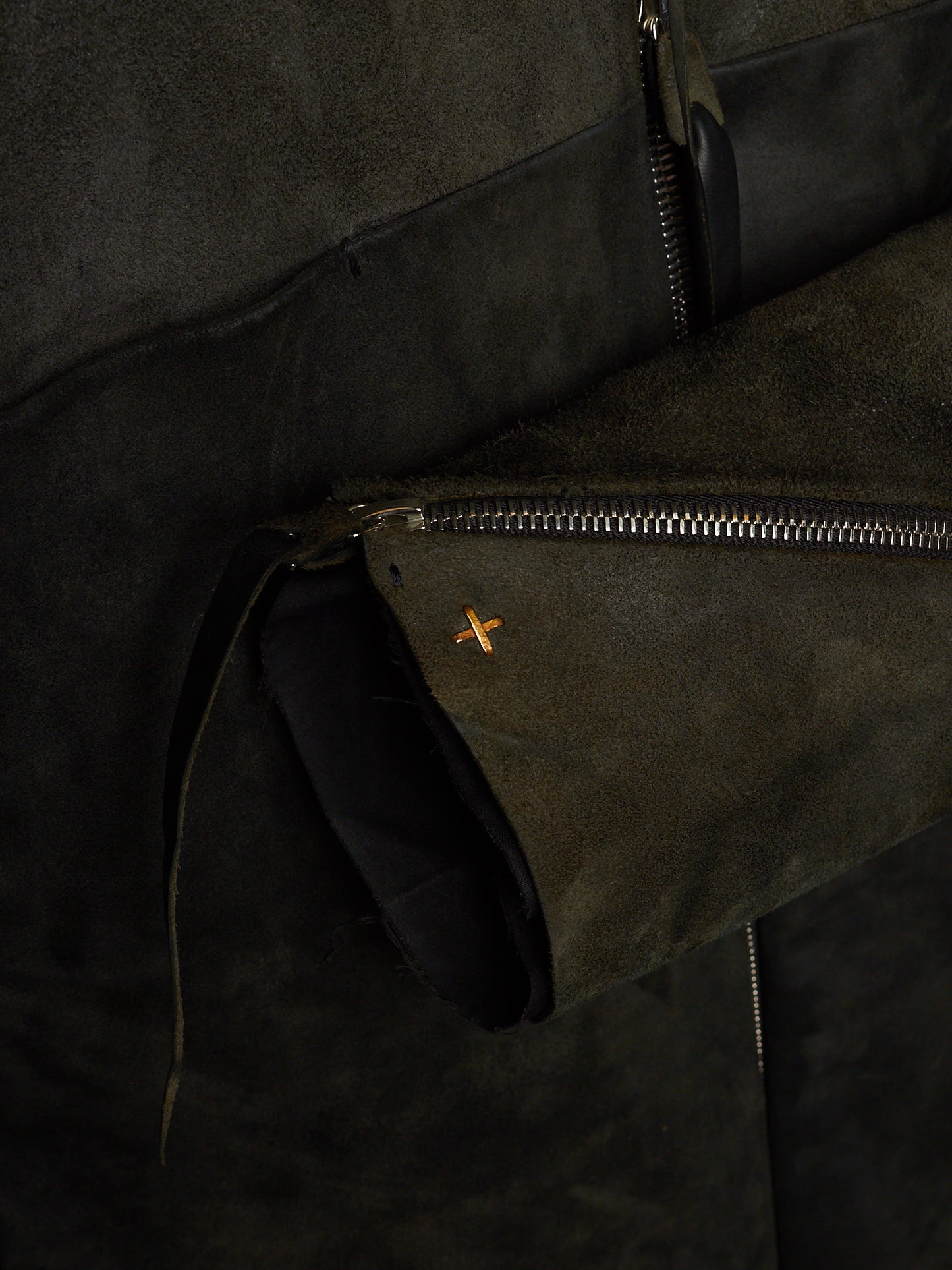 Close-up of a dark olive green suede jacket with a silver zipper and a small orange embroidered cross logo.